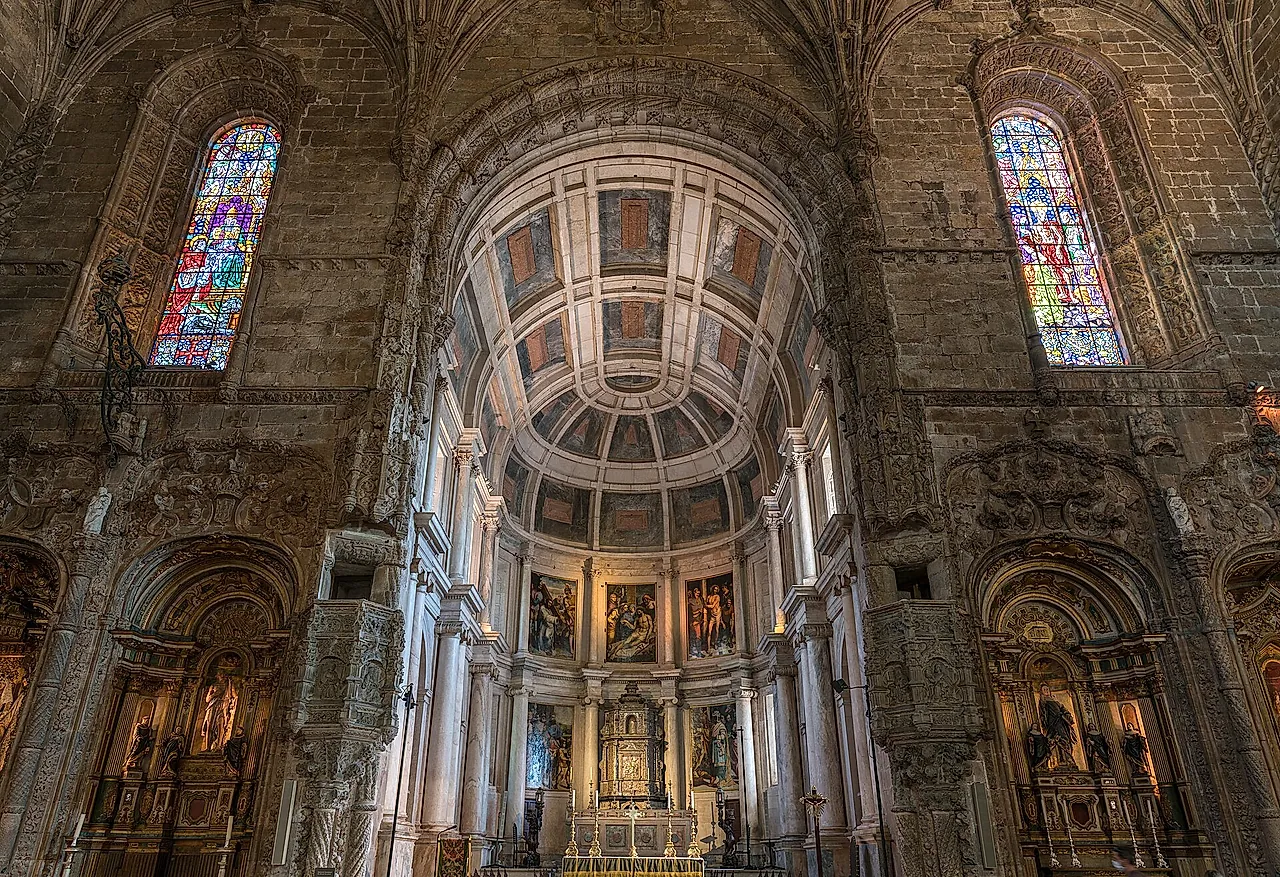 The apse of the church seen from the nave: a coffered barrel-vaulted chancel with paintings and an altar, stained-glass windows on either side and side chapels glowing in warm light
