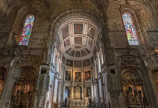 The apse of the church seen from the nave: a coffered barrel-vaulted chancel with paintings and an altar, stained-glass windows on either side and side chapels glowing in warm light