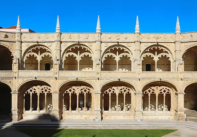 Two storeys of carved stone arcades around the cloister courtyard under a clear blue sky, with slim pinnacles along the roofline and a strip of lawn in front