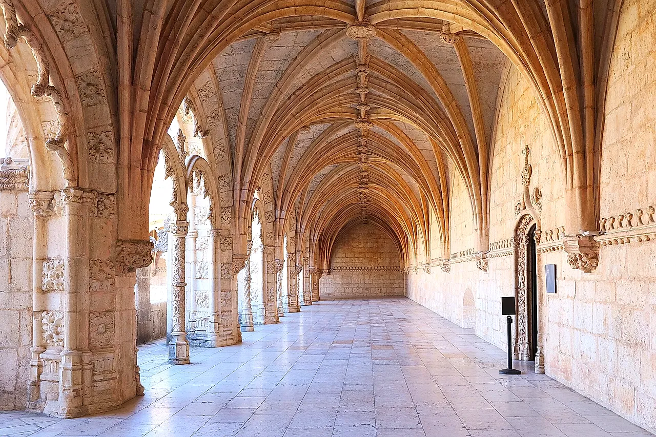 A long vaulted gallery of the cloister, with a ribbed stone ceiling, carved pillars on the left and a pale stone floor leading to the far end