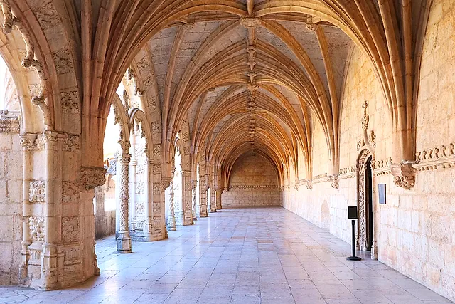 A long vaulted gallery of the cloister, with a ribbed stone ceiling, carved pillars on the left and a pale stone floor leading to the far end