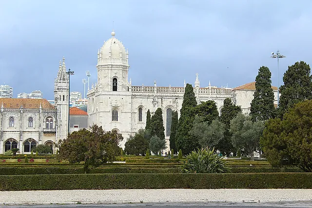 The monastery seen across the clipped hedges of the Praça do Império garden, with the domed bell tower and the church in pale stone among tall cypress trees