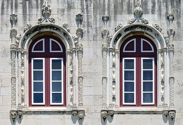 Two arched windows with dark red frames set in a pale stone wall and surrounded by elaborately carved twisted columns and stone ornament