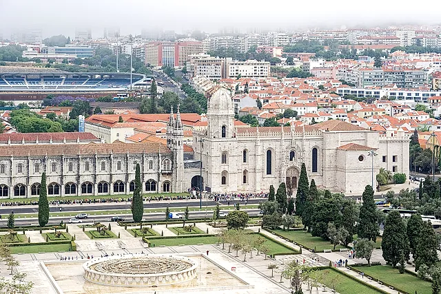 Aerial view of the long monastery façade beside the gardens, with the church at the right, the red roofs of Lisbon and a stadium in the hazy distance