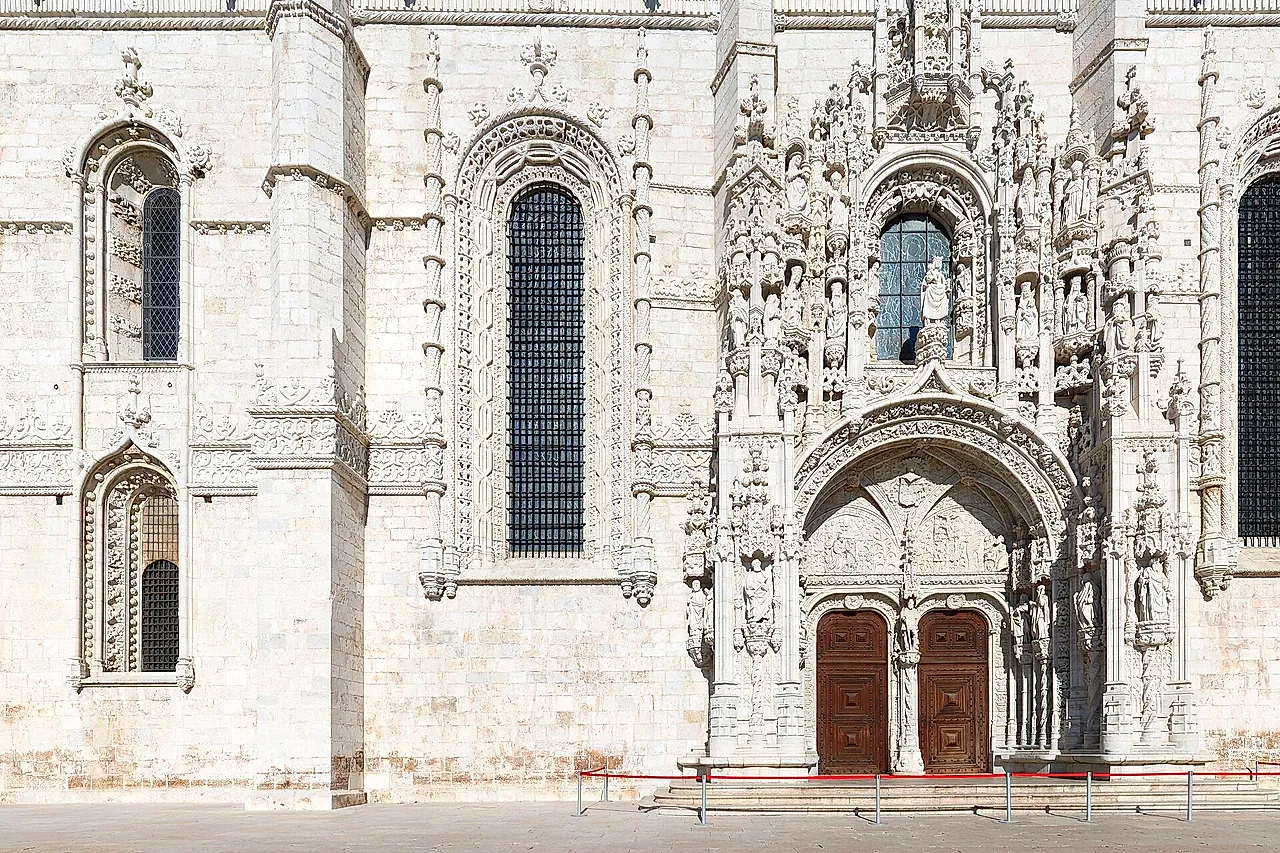 The richly carved south portal of the monastery church, with rows of sculpted figures around the arch above two dark wooden doors, beside a tall arched window in the pale stone wall