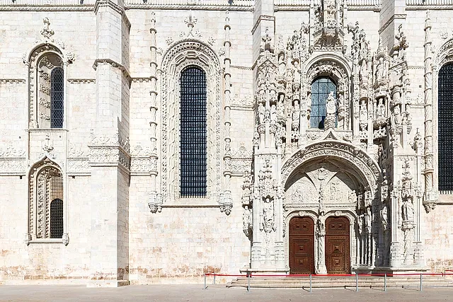 The richly carved south portal of the monastery church, with rows of sculpted figures around the arch above two dark wooden doors, beside a tall arched window in the pale stone wall