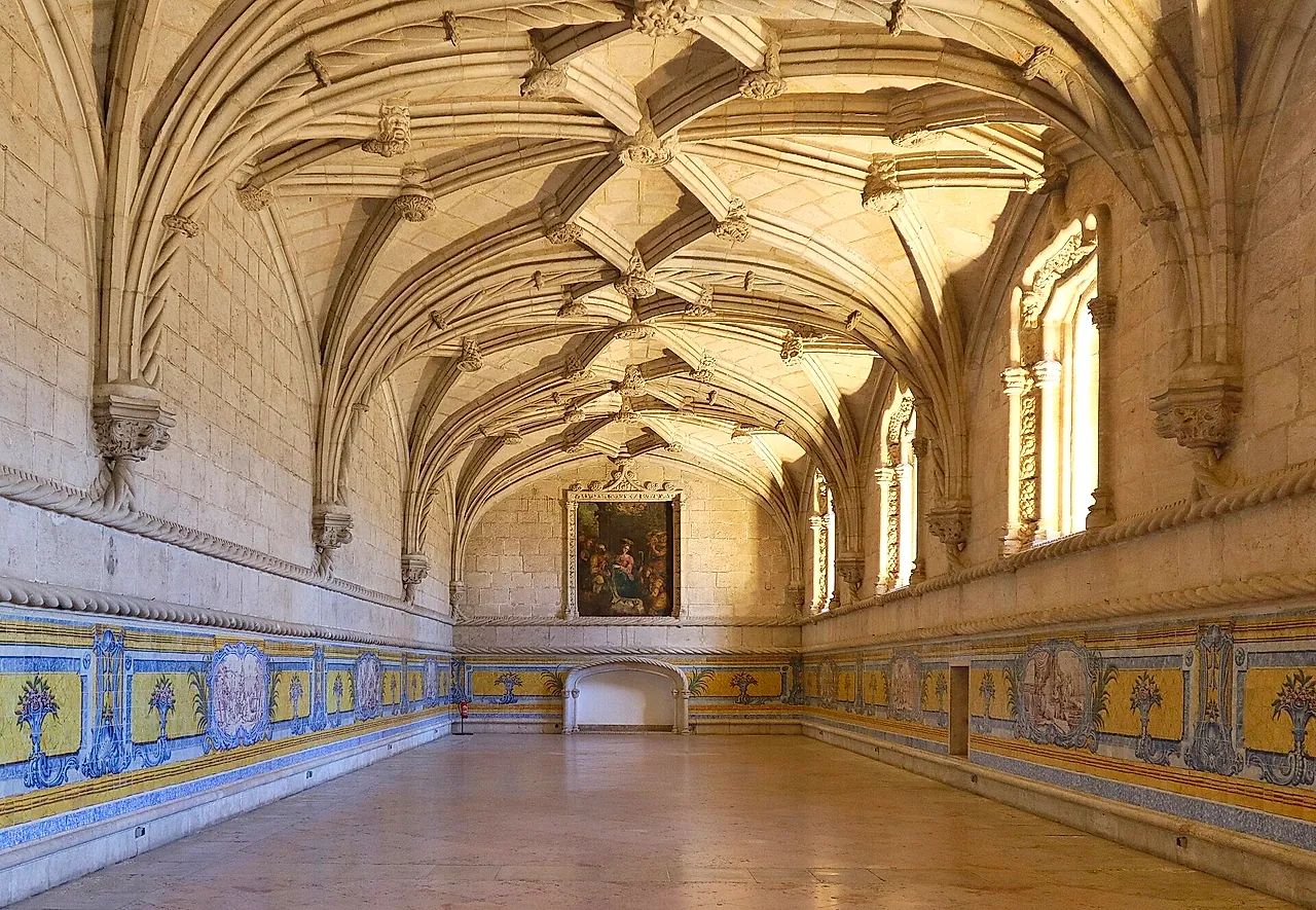 The long refectory hall with a ribbed vault, blue and yellow tile panels along the lower walls and a painting at the far end, lit by sunlight from the windows
