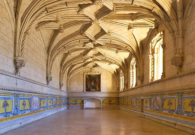 The long refectory hall with a ribbed vault, blue and yellow tile panels along the lower walls and a painting at the far end, lit by sunlight from the windows