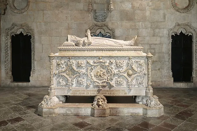 A white marble tomb carved with a ship and leafy ornament, topped by a reclining figure and resting on carved lions, between two dark pointed recesses on a stone floor
