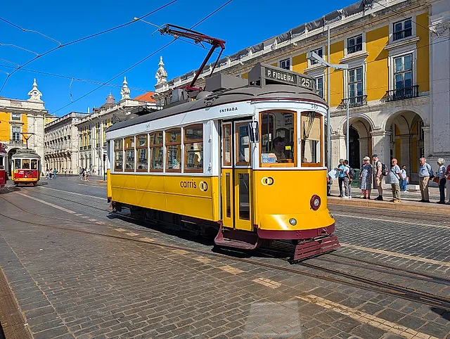 A yellow and white vintage Lisbon tram on the cobbled square of Praça do Comércio, with the yellow arcaded buildings behind it and a few people waiting at the stop, under a clear blue sky