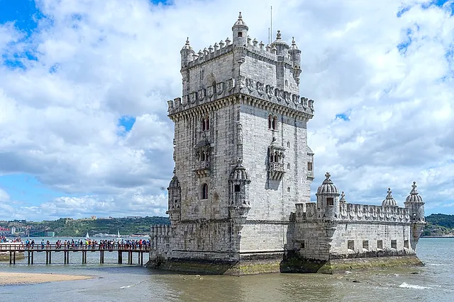 Belém Tower seen from the river side under a sky of white clouds, with its decorated turrets and a walkway with visitors at the left