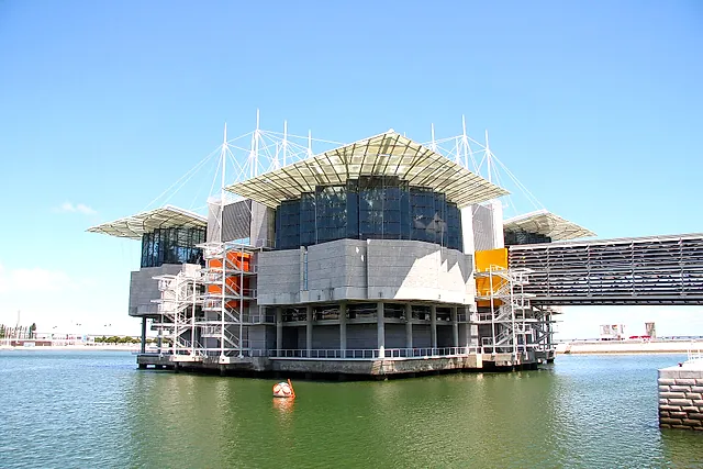 The Lisbon Oceanarium building rising from the water of the dock, with glass canopies, white masts and cables above grey concrete walls, a footbridge on the right and a round mascot buoy in the foreground, under a blue sky