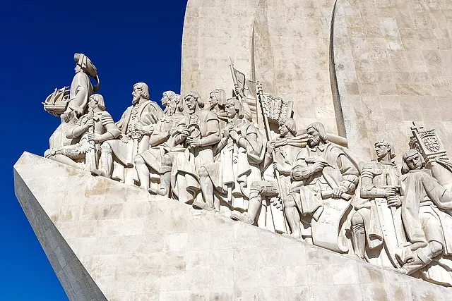 The sculpted prow of the Monument to the Discoveries, with a row of stone figures led by one holding a small ship, against a deep blue sky