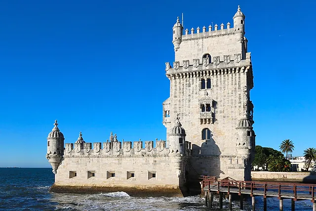 Belém Tower, the pale stone tower with turrets and a low bastion, standing at the water's edge with a wooden walkway under a blue sky